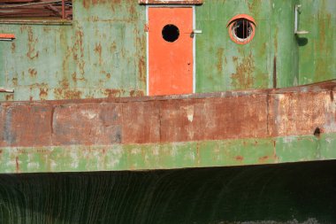 Yıpranmış yeşil bir sanayi gemisinin güverte boyunca kırık bir pencere ve ağır pas dokuları ve gövdesi olan canlı turuncu bir kapısı vardır.