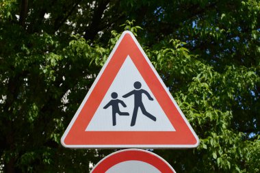 Çocukların karşıya geçtiğini gösteren üçgen bir yol işareti parlak gün ışığı altında yemyeşil ağaç yapraklarının önünde konumlandırılmıştır.