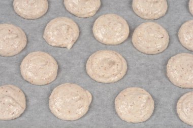 Fırınlanmamış birkaç sıra bej fransız badem kurabiyesinin yüksek açılı görüntüsü beyaz parşömen kağıdına konmuş hamur hamuru parçaları.