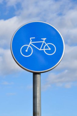 Beyaz bisiklet sembollü dairesel mavi trafik levhası yumuşak beyaz bulutlu mavi gökyüzüne karşı metal bir direğin üzerinde duruyor.