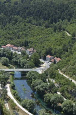 Tarihi Knin Kalesi 'nden yüksek açılı bir manzara, köprünün altından akan açık mavi Krka nehrinin geleneksel evlerin ve yemyeşil bir ormanın yanında olduğunu gösteriyor.