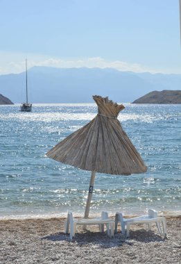 Krk adasındaki Vela Luka 'da çakıl taşlı bir plajda, mavi denizde yelkenli, Velebit Dağı' nda hasır güneş şemsiyesi ve şezlonglar.
