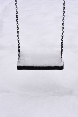 Soğuk bir kış günü, beyaz bir kar tabakasıyla kaplı metal zincirlerden sallanan boş bir siyah oyun alanı.