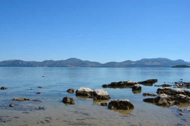 A serene landscape showing the rocky shore of Drace on Peljesac peninsula looking across the Malo More towards the mountains of mainland Croatia under a clear sky