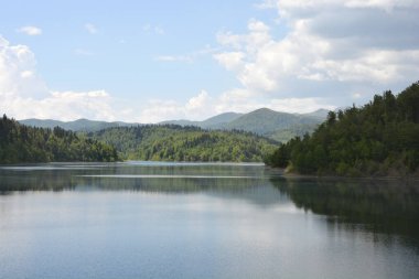 Gorski Kotar 'daki Lokve Gölü' nün sakin suları yoğun yeşil tepeler ve bulutlu mavi gökyüzünün altındaki ormanlarla çevrili.