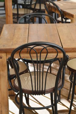 Dikey çekimde birkaç tahta masa ve hasır koltuklu siyah metal sandalyeler gün boyunca açık bir kafe ortamında sergileniyor..