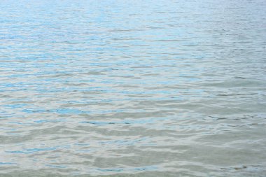 Sakin mavi denizin yüzeyinde, berrak bir gün ışığı altında hafif dalgalar ve yumuşak yansımalar vardır.