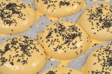 Ham hamur çöreklerinin yüksek açılı görüntüsü yumurta yıkama ile kaplanmış ve üzerine siyah susam tohumları konmuş. Pişirmeden önce beyaz ekmek kağıdına yerleştirilmiş.