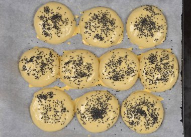 Siyah susam tohumlarıyla kaplı pişmemiş hamburger çörekleri ve ahşap bir masaya yerleştirilmiş parşömen kağıdındaki yumurta yıkama tepsisinin üst görüntüsü.