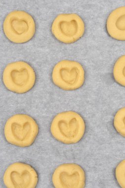 Pişmemiş yuvarlak kurabiyelerin yukarıdan görünüşü kalp şeklinde girintiler beyaz parşömen kağıdına yerleştirilmiş fırın için hazır siyah bir yemek tepsisi.