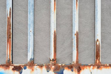 Gri desenli düz bir yüzeye karşı görünen turuncu pas lekeleri olan dikey beyaz metal çubukları kapat