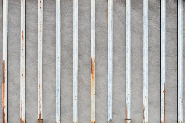 Görünür pas lekeleri ve aşınması olan birden fazla dikey beyaz metal çubuğun düz bir görünümü var.