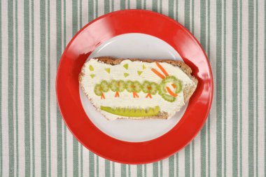 Krem peynirli yaratıcı bir sandviç ve çizgili masa örtüsüne yerleştirilmiş kırmızı ve beyaz bir tabakta sebze tırtılı.