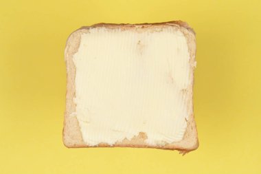 Üzerinde kremalı tereyağı katmanı olan tek bir dilim beyaz ekmeğin üst görüntüsü sarı bir arkaplana yayılmış.