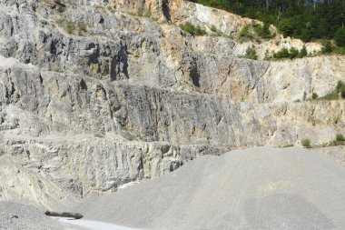 Kayalık kayalıklara ve yeşil bir ormana karşı ön planda ezilmiş büyük bir çakıl yığınının yer aldığı aktif bir taş ocağının geniş bir görüntüsü.
