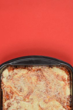 Fırınlanmamış lazanya tabakalarının yüksek açılı görüntüsü et soslu ve bechamel üzerine siyah bir tavada dilimlenmiş peynirle parlak kırmızı bir arka planda.