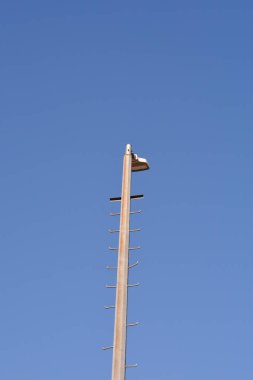 Metal tırmanma merdivenleri ve üzerinde tek bir lamba bulunan uzun beton bir sokak lambası açık mavi gökyüzü arka planında duruyor.