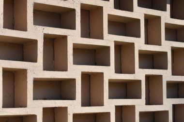 Bej beton bir duvarın köşegen perspektifi, köşeli oyukların ve dekoratif esinti bloklarının tekrarlayan geometrik desenini içerir.