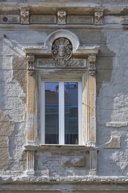 Klasik beyaz bir pencere, kadın yüzü ve çiçek süslemeleri içeren yıpranmış taş bir cepheye kurulur.