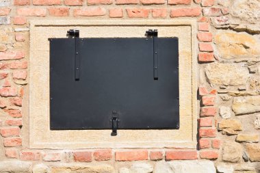 Eski kırmızı tuğlalar ve doğal açık renkli taşlardan oluşan tarihi bir bina duvarına siyah metal bir kepenk monte edilmiştir.