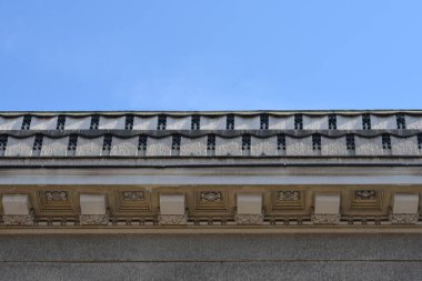 Detaylı taş oymaları ve dekoratif desenler, klasik bir binanın üst köşesini ve çatısını açık mavi gökyüzüne karşı süsler.