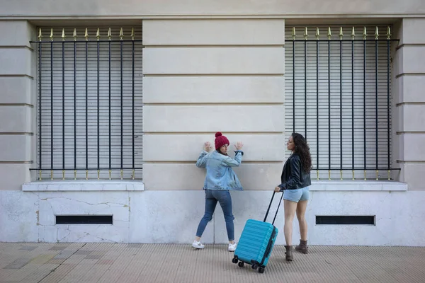 Amici donne in piedi sulla strada di fronte al muro con una valigia Immagine Stock