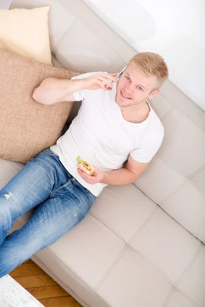 年轻男子坐在沙发上吃三明治 — 图库照片