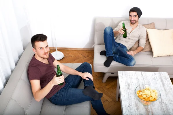 Два друга с пивом и чипсами в гостиной — стоковое фото