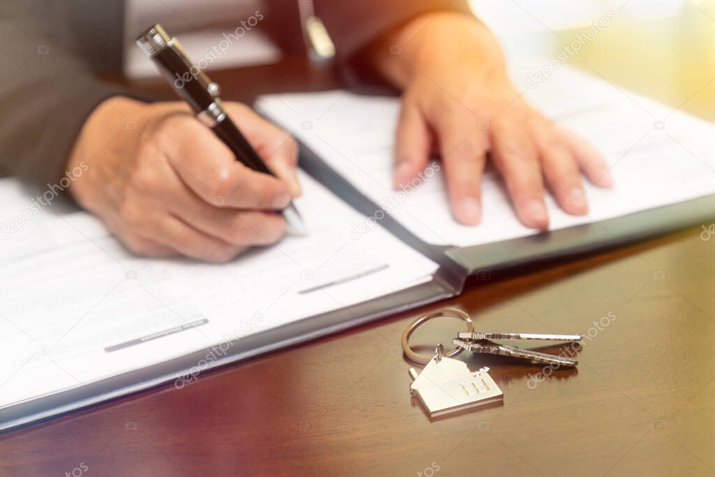 Woman signing real estate contract papers with house keys and home keychain in front.