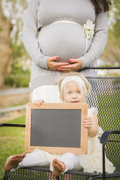 Maman enceinte derrière bébé fille dans chaise tenant tableau noir vierge — Photo