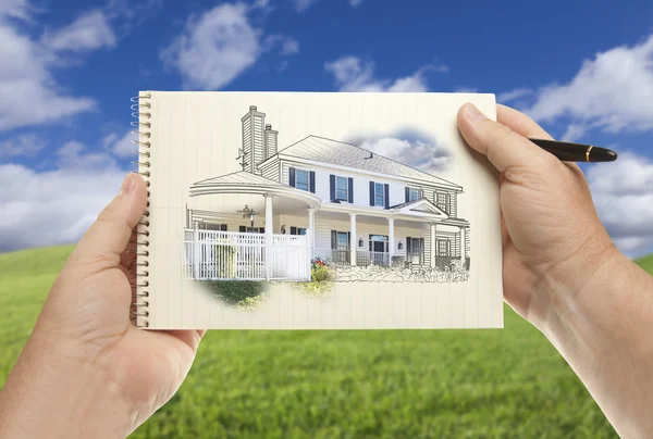 Mãos segurando papel com desenho da casa sobre campo de grama vazio — Fotografia de Stock