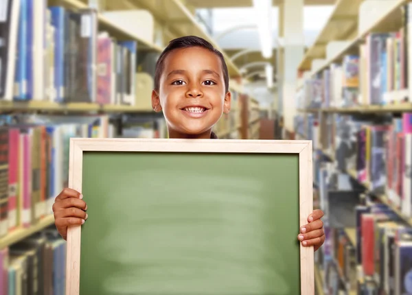 Un garçon hispanique souriant tenant un tableau de craie vide à la bibliothèque — Photo