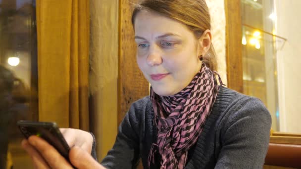 Mujer joven que utiliza la aplicación en un teléfono y beber café y sonreír en la cafetería. 4K UHD — Vídeo de stock