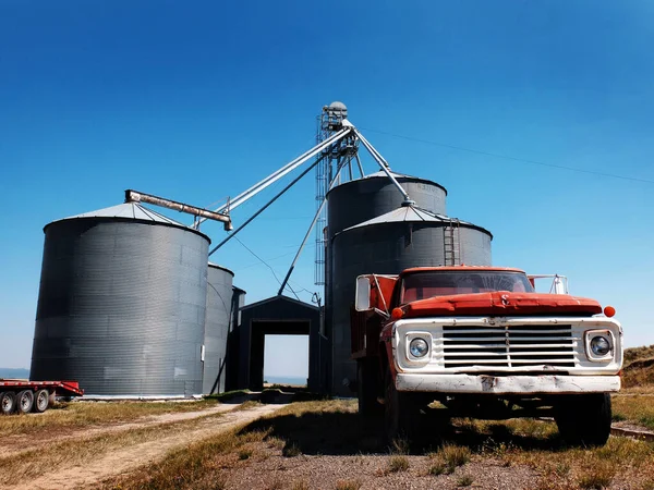 Old red work truck parked in front of several large steel grain silos on farm