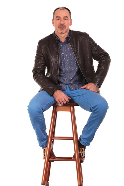 Homem de jaqueta de couro posando no banquinho com as pernas abertas — Fotografia de Stock