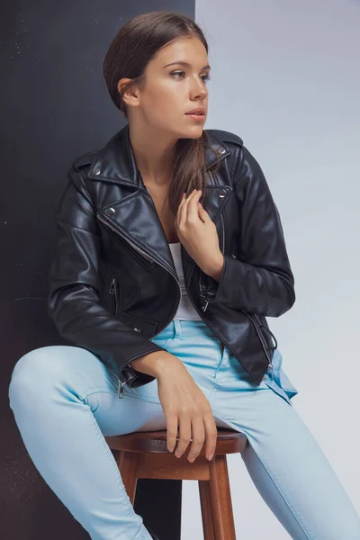 Thinking fashion model adjusting her leather jacket and looking away while sitting on a stool on black and white studio background