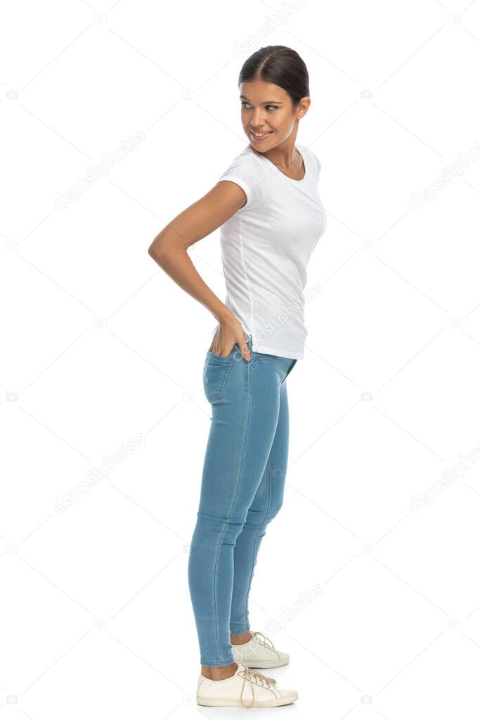 Side view of happy casual woman looking over shoulder with hand in pocket, standing on white studio background