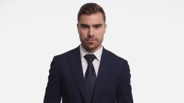 Mutlu genç işadamı gülümsüyor, lacivert takımını ilikliyor ve başparmak işareti yapıyor. Stüdyoda beyaz arka planda tek başına duruyor.