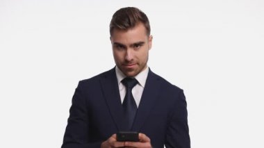 Lacivert takım elbiseli genç işadamı elinde cep telefonu, mesaj atıyor, gülümsüyor ve başını sallıyor, beyaz arka planda tek başına duruyor.