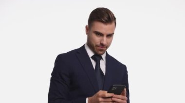 Lacivert takım elbise giyen, cep telefonu tutan, e-posta okuyan, gülümseyen, kollarını kaldırıp tezahürat yapan, stüdyoda beyaz arka planı işaret eden mutlu genç adam.