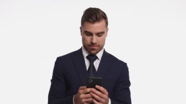 Lacivert takımlı çekici bir adam elinde telefonla e-postaları okuyor, parmaklarıyla işaret ediyor ve gülümsüyor. Stüdyoda beyaz arka planda tek başına duruyor.