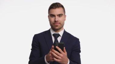 Lacivert takım elbiseli mutlu genç işadamı, elinde telefon, mesajlar okuyor, gülümsüyor ve dans ediyor. Stüdyoda beyaz arka planda izole halde.