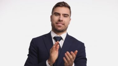Lacivert takım elbiseli gururlu genç adam başını sallıyor ve mutlu bir şekilde alkışlıyor, parmakla işaret ediyor ve gülümsüyor, stüdyoda beyaz arka planda tek başına duruyor.