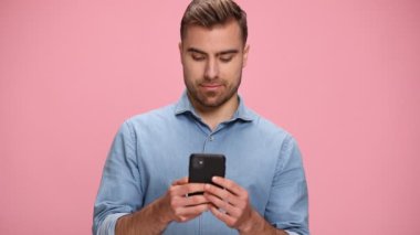 happy casual man in blue denim shirt holding phone, reading messages and making thumbs up gesture on pink background in studio