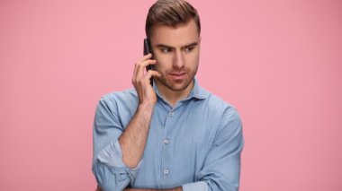 young casual guy in blue denim shirt talking on the phone, nodding and smiling, standing on pink background in studio
