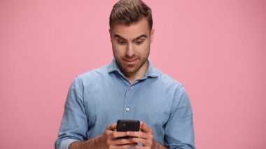 amazed young man wearing denim shirt, reading messages, holding fist up and cheering victory, laughing on pink background in studio