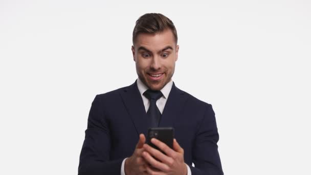 ネイビーブルーのスーツを着た若い実業家が電話をかけ驚きのメールを読んで拳を握り勝利を祝い笑顔で白い背景に孤立して立っている — ストック動画