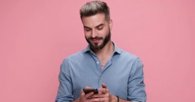 young casual man writing an email on his cell phone, giving a thumbs up and feeling excited on pink background