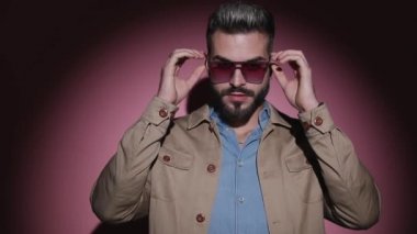Kot gömlekli havalı adam bej ceketini ayarlıyor, gözlüklerini çıkarıyor ve poz veriyor, stüdyoda pembe arka planda güven içinde kollarını kavuşturuyor.