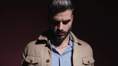 Kot gömlekli gizemli genç adam aşağıya bakıyor, duygusal olarak gözlük takıyor ve stüdyoda pembe arka planda moda ışığı altında poz veriyor.